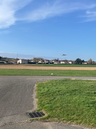 The image captures an outdoor scene under a clear blue sky with minimal scattered white clouds, taken from a ground-level perspective at the Aerodrome de Vienne Reventin in Chonas-l'Amballan, France. In the upper mid-ground, a single-engine, high-wing light aircraft, colored red, is in flight, positioned above a distant residential area. This area consists of multiple houses with light-toned facades and sloped, tiled roofs, intermingled with trees, some bare and others retaining foliage. Beyond the houses, distant hills are vaguely visible. Below the residential zone, the landscape transitions to agricultural fields, displaying patches of vibrant green grass, brown tilled earth, and lighter green cultivated sections. The immediate foreground features a dark gray asphalt surface, showing minor cracks, with a black rectangular metal drain grate embedded in the bottom-left corner. This asphalt surface is bordered on its right by an expanse of bright green grass. A small, yellow conical marker is present on the edge of the green field in the mid-ground, slightly right of center. No individuals are present in the frame.