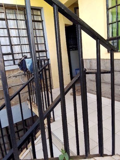 The image depicts a multi-story building's stairwell and landing area in Kalimoni ward, Kenya. A black metal railing with vertical bars and a handrail descends diagonally from the foreground left to the mid-ground right. The walls are painted a cream or light yellow, with a lower section constructed from grey concrete blocks. On the left, a window with black metal security bars is visible, covered by white curtains. Clothes, including blue and dark garments, are hanging on a thin line or wire tied to the security bars of this window. To the right, a second window with black metal security bars features green-tinted glass. The landing floor is covered with light-colored, possibly white or cream, square tiles laid in a diagonal pattern. A pair of light-brown woven sandals rests on the tiled floor to the right of the center. In the background, a dark, open doorway leads into a shadowed interior, where a partial view of a blue object, possibly a chair or stool, is visible. A small green plant with a few leaves grows from the base of the railing in the lower center foreground. No individuals are present in the scene.