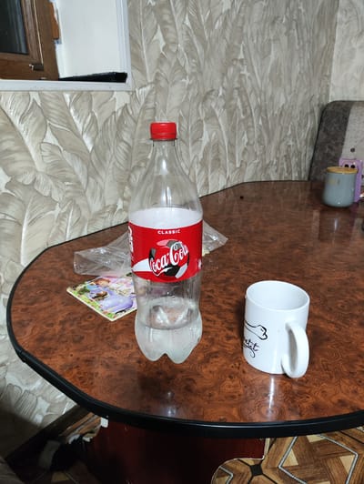 A partially filled bottle of Coca-Cola Classic stands on a dark, wood-grained, circular table. To the left of the bottle is a crumpled plastic bag. In front of the bottle is a colorful coaster featuring the character Sofia the First. To the right of the bottle is a white ceramic mug with a black outline drawing of a cat and the word "petit" printed on it. In the background, a textured wallpaper with a leaf pattern is visible. A portion of a striped armchair is also visible to the right. The lighting in the image appears to be artificial and comes from above. The scene depicts a casual setting, likely indoors, on a table.