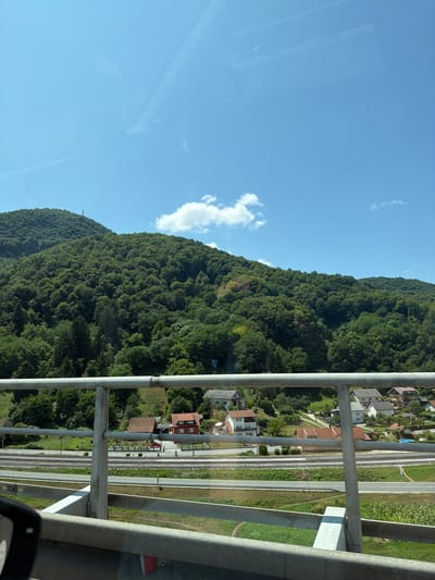 The image captures a view from a railing overlooking a rural landscape in Ravninsko, Croatia. The foreground features a metal railing and a portion of a car's side mirror. Beyond the railing, a road and train tracks are visible, separating the foreground from a village with several houses. In the background, a lush, tree-covered mountain dominates the scene under a bright blue sky with scattered white clouds.