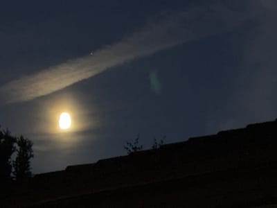 The image depicts a night sky over a dark, silhouetted roofline, likely in Neumünster, Germany. In the upper left quadrant, a bright, luminous moon is partially obscured by wispy clouds, casting a soft glow. To the upper right of the moon, a faint, elongated cloud formation stretches diagonally across the sky. A small, bright point of light, possibly a star or a distant light source, is visible within this cloud. In the mid-right portion of the sky, a nebulous, greenish cloud is present. The foreground is dominated by the dark, textured surface of a tiled roof, with silhouetted tree branches visible against the sky on the left side. The overall scene is one of a nocturnal urban or suburban landscape.