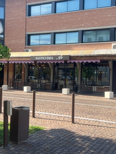 This is a vertical, eye-level shot of a building facade with a pastry shop. The building is made of reddish-brown brick with large, dark-framed windows. The shop, "PASTICCERIA da Filippo," has a yellow awning and a glass storefront. The entrance is marked by two sets of glass doors. Inside, the shop is lit with warm, ambient light, and trees are visible through the glass, suggesting an outdoor seating area or a view of greenery.
The scene is outdoors on a bright, sunny day, indicated by the strong shadows cast on the cobblestone pavement and the bright lighting. The pavement is laid in a diamond pattern, typical of many Italian streets and piazzas. There are several brown metal bollards and a dark brown trash receptacle visible along the walkway. The overall atmosphere is that of a peaceful, ordinary day in a European town.
There is no visible activity or event taking place; the scene appears static, focusing on the architecture and the shop's exterior. The time of day is likely midday, given the angle of the shadows. The location is identified as Campodarsego, Italy, which is further supported by the architectural style and paving. The text "PASTICCERIA da Filippo" is clearly visible on a dark sign above the shop's entrance.