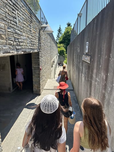 The image shows a view from above, looking down on a group of people walking along a narrow, walled pathway. The pathway appears to be constructed from stone on one side and concrete on the other. Some of the people are walking up a set of stairs. One person is entering a small, enclosed structure. The location is Orta San Giulio, Italy.
