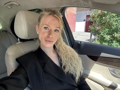 A blonde woman with blonde curly hair, wearing a black blazer, sits in the driver's seat of a car. She is looking directly at the camera with a neutral expression. The interior of the car is visible, with light-colored leather seats and dark wood trim on the door. Outside the car, a brick building and a lush green bush are visible. The scene appears to be in the city of Kommolovo, Russia, as indicated by a sticker on the car's window.