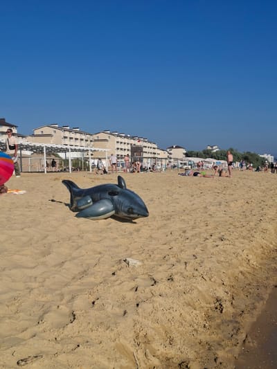The image depicts a sandy beach with several people present. An inflatable shark toy lies on the sand in the foreground. In the background, there are buildings lining the beach, suggesting a resort or populated coastal area. Some people are lying on the sand, while others appear to be walking or standing. The sky is clear and blue, indicating fair weather, likely on a sunny day. The overall scene suggests a casual beach outing or vacation. There is no visible text in the image. The orientation of the image is rotated.