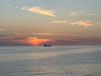 The image depicts a cargo ship sailing on a calm sea during sunset. The sky is a gradient of colors, with hues of orange, pink, and blue, indicative of the sun setting below the horizon. Wispy clouds are scattered across the sky, catching the warm light of the setting sun. The sea is smooth with gentle ripples, reflecting the colors of the sky. The cargo ship, a dark silhouette against the vibrant sky, is positioned slightly to the left of the center of the frame. The scene is serene and tranquil, capturing a moment of quiet passage at sea. The location is not specified in the image metadata.