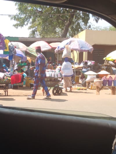 The image captures an active outdoor market scene in Omupo, Nigeria, observed from inside a vehicle.
In the foreground, a male figure walks from left to right, wearing a red cap and a blue and red patterned long-sleeved shirt and matching trousers, with light-colored sandals. He is positioned near a two-wheeled utility cart laden with small packaged items. To his right, a female figure stands facing away, balancing a large white bag on her head. She wears a white long-sleeved shirt with "AL-HUDAA" printed in blue text on the back and a patterned skirt, standing beneath a striped market umbrella providing shade.
The market extends into the mid-ground, characterized by numerous stalls, many shaded by colorful market umbrellas and some with corrugated metal roofs. Merchandise includes various hanging clothing items on the left, small packaged goods on tables, and produce such as red onions displayed in purple plastic baskets, alongside sacks of bulk items, on the right. Multiple other individuals are visible throughout the scene, some seated, others standing, suggesting vendors and shoppers engaged in commerce.
Environmental details include an unpaved or dirt road/pathway, distinct shadows cast by strong sunlight, and a large tree with green foliage visible in the upper background against a clear sky. A light brown building with a dark opening is partially visible on the far right mid-ground. The dark interior of a vehicle dashboard occupies the bottom portion of the frame.