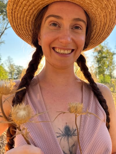 A woman wearing a straw hat and a pink textured tank top smiles at the camera. She has dark brown hair in two braids, one on each side of her head. In front of her chest, she holds a dried thistle flower. A tattoo of a thistle is visible on her chest. The background consists of blurred greenery and a bright blue sky, suggesting an outdoor setting in Katzrin, Israel.