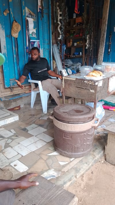 A man is sitting on a plastic chair in front of a shop in Asubi, Nigeria. The shop's facade is primarily blue, with shelves displaying various items, including tools, pipes, and hardware. The man is wearing a black T-shirt and khaki pants. He is holding a small, round object. There is a table next to him with food in containers. A large brown cooler with the word "CUTIE" on it sits in the foreground. The floor is a mix of tile and exposed earth. Another person's hand is visible in the lower left corner of the frame.