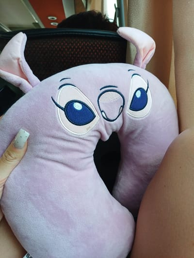 A close-up shot focuses on a pink, plush neck pillow shaped like a character with large blue eyes and pink ears, positioned on a person's lap. The pillow is adorned with embroidered facial features, including eyebrows, eyelashes, and a small mouth. A hand with a nail adorned with rhinestones is visible on the left side of the frame, touching the pillow. The background is blurred, showing hints of a car interior, including seats and a window with curtains. No specific location within Lumina, Romania, is discernible from the image.