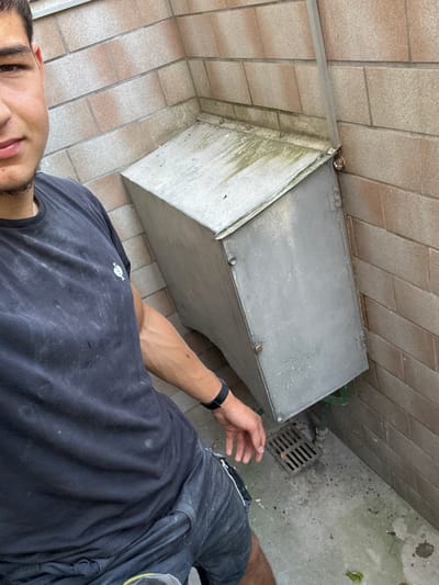 A young man stands in front of a metallic box attached to a brick wall. The man is wearing a dark t-shirt and shorts, and his face is partially visible. The metallic box appears to be an outdoor electrical enclosure or utility box. It is situated in an outdoor setting, with a concrete ground and a drain grate visible. The brick wall suggests an urban or suburban environment. The scene does not provide any specific clues to pinpoint its location within Brühl, Germany.