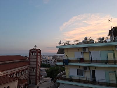 The image shows a city skyline at dusk, with a church on the left and apartment buildings on the right. The sky is a gradient of pale blue and pink, with wispy clouds.

The church features a red-tiled roof and a bell tower with alternating red and white stone bands. A cross tops the tower. The church's facade is also adorned with red and white banding and arched windows.

To the right, a multi-story apartment building is visible, painted in shades of yellow and green. It has balconies with railings, and several air conditioning units are mounted on the exterior walls. A pair of blue jeans is hanging from a clothesline on one of the balconies. Another apartment building with similar features is partially visible in the background.

In the distance, other buildings, trees, and a road with a car can be seen, indicating an urban environment. The overall scene suggests a residential area within the city of Stavroupoli Municipal Unit, Greece.