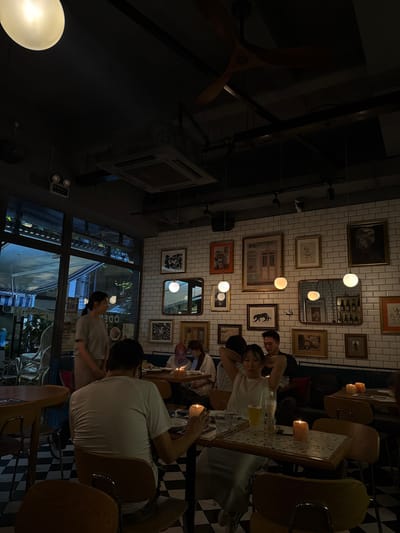 The image depicts the interior of a dimly lit restaurant in Xuhui District, China. The walls are tiled with white subway tiles, and adorned with numerous framed artworks and mirrors. Several round pendant lights and a ceiling fan are visible. Tables are set with candles, creating a warm ambiance. People are seated at various tables, engaged in conversation or dining. One woman stands near a large window looking out, while others are seated at tables. The flooring is a black and white checkered pattern. The overall atmosphere suggests a casual dining or social gathering.