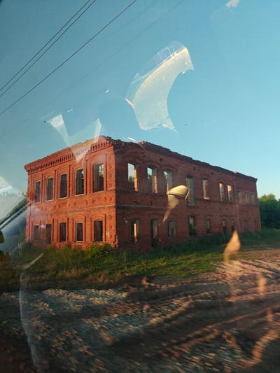 The image captures an exterior view of a dilapidated, L-shaped, two-story red brick building under a clear blue sky in Nikolo-Beryozovka, Russia. The building, likely an abandoned industrial or residential structure, exhibits significant deterioration, with numerous empty window frames and an incomplete or absent roof structure. It is situated on an uneven landscape featuring patches of green grass and a dirt path or road in the foreground. Green foliage and trees are visible to the right and behind the structure. The scene is illuminated by warm, golden sunlight, casting shadows across the building's facade and the ground.
The photograph was taken through a window, resulting in prominent reflections and distortions across the entire frame. In the upper left quadrant, several parallel dark lines, resembling power lines or wires, are reflected against the sky. The upper half of the image contains translucent reflections of interior elements, including curved, lighter shapes that suggest window frames, vehicle pillars, or interior structures, and a darker, rounded silhouette possibly indicating a person's head or shoulder. Small, dark, bird-like specks are visible in the sky. Streaks of light and glare are present, particularly in the lower right corner and integrated within the reflections, indicating light interacting with the glass surface. The overall composition suggests a fleeting view from a moving vehicle.
