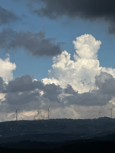 The image depicts a hilly landscape under a dramatic sky. Several wind turbines are visible along the ridge of a dark green, vegetated hill, silhouetted against a backdrop of clouds. The sky is a mix of deep blue, dark gray storm clouds, and bright white cumulus clouds, with the sun illuminating some of the cloud formations. The foreground is a dark, indistinct mass of land, possibly a lower hill or valley. The scene suggests a location with renewable energy infrastructure in a natural environment. The specific location in the city of Đa Lộc, Vietnam is not discernible from the image itself. There are no people or discernible actions taking place.