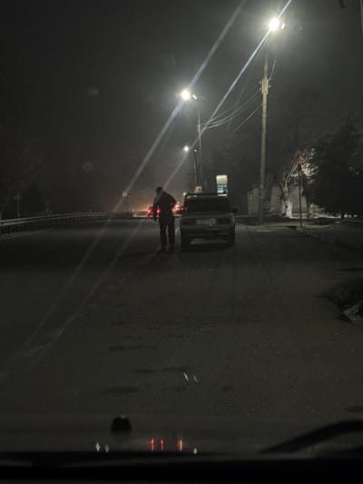 A street scene at night features a dark vehicle parked on the right side of the road. A person stands near the vehicle. Streetlights illuminate the scene, casting long, bright beams through the foggy or hazy air. The road is dark and wet, reflecting the lights. There are buildings in the background. The view appears to be taken from another vehicle with a dark window frame on the right side of the image.