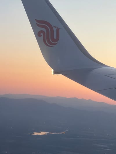 The image shows an airplane wing with the Air China logo on its vertical stabilizer. Outside the window, a hazy view reveals mountains and water, possibly at sunset. The scene is likely taken during a flight near Shunyi District, China, considering Air China's major hub is Beijing Capital International Airport, located in that area.