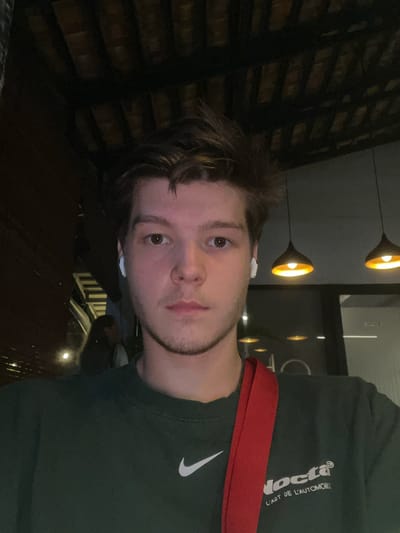 A medium indoor shot presents a young, fair-skinned man with brown hair, a light beard, and brown eyes. He is wearing white wireless earbuds and an olive-green t-shirt with a white logo and a red strap across his shoulder. Behind him, the ceiling features dark wooden latticework. In the background, there are multiple globe-shaped hanging light fixtures. A window or doorway frames a dimly lit space with additional people and what appears to be a parked car. The lighting suggests it may be evening. While the prompt specifies Benoa, Indonesia, there's no visual information to confirm the location.