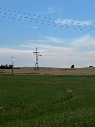The image depicts an open, rural landscape under a clear blue sky with scattered white clouds. In the foreground, a vibrant green grassy field stretches across the bottom of the frame. Beyond this field, a golden-brown expanse of a crop, likely wheat or barley, dominates the mid-ground. Two prominent electricity pylons stand tall in the mid-ground, one closer and larger than the other in the distance. These pylons are connected by numerous overhead power lines that traverse the frame horizontally, extending towards the horizon. Several smaller, dark green trees are visible on the far left of the mid-ground, near the base of the leftmost pylon. In the far distance, a lone dark green tree stands out against the horizon, and a cluster of trees forms a dark silhouette on the right. The overall environment suggests a rural setting, possibly in the vicinity of Rudelzhausen, Germany, characterized by agricultural fields and electrical infrastructure. There are no people or animals visible in the image.