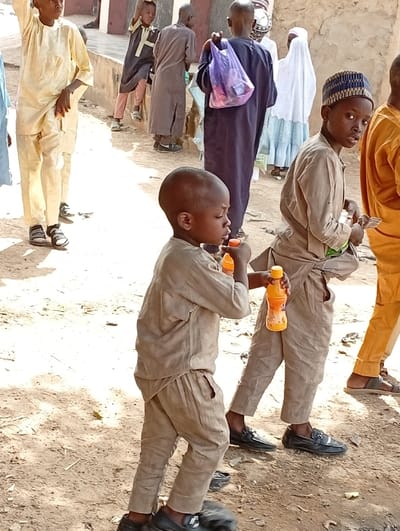 A group of people stand outside. Several are children, dressed in traditional clothing. One child in the foreground drinks from a bottle. Another child wearing a kufi stands beside him, looking toward the camera. People in the background include adults wearing robes and head coverings. The ground is dusty, and a building can be seen in the background.