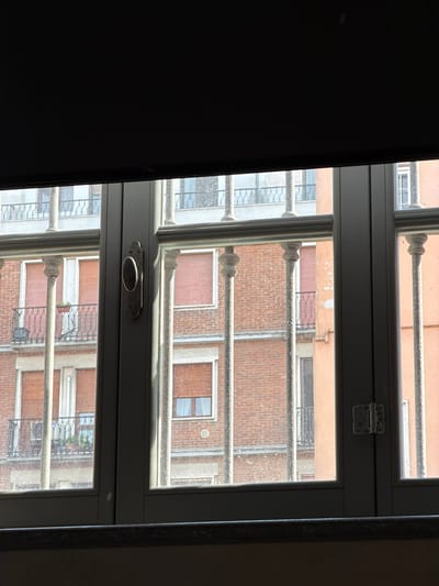 This image depicts a view through a window with a roller blind partially drawn, revealing a sunlit urban scene. The window has a dark frame and vertical bars in front of it. Beyond the window, multiple buildings with brick facades and architectural details like balconies and shutters are visible. Sunlight highlights some of the windows and architectural elements, creating a play of light and shadow. The environment appears to be a city, possibly Piacenza, Italy, given the architectural style, though no specific landmarks are identifiable to confirm the location. There are no people visible in the image. The scene is static and observational.