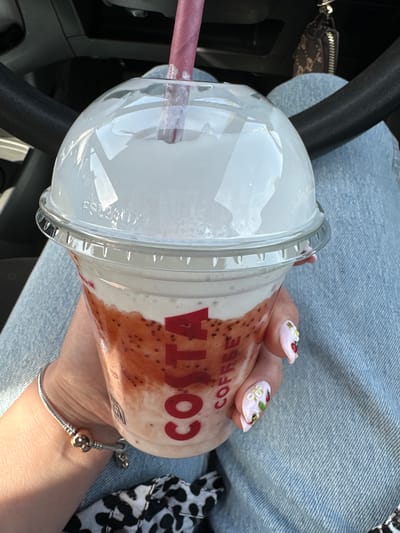 A person's hand, adorned with a silver bracelet and decorated nails featuring floral and insect designs, holds a clear plastic cup filled with a strawberry-flavored drink and topped with whipped cream. The cup has the "COSTA COFFEE" logo printed on it in red. A pink paper straw extends from the domed lid. The background shows the interior of a vehicle, including the steering wheel and dashboard, and the person's light blue denim jeans. The scene is lit by natural sunlight, suggesting it is daytime. The location is the city of Carlow, Ireland.