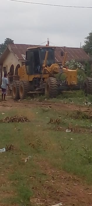 The image displays an outdoor scene in the city of Ijotan, Nigeria, under an overcast sky. A large, yellow motor grader is centrally positioned, appearing stationary, with its blade assembly visible towards the front and its six prominent tires on a dirt and grass surface. To the left of the grader, a single individual is standing, facing partially right, wearing a light blue short-sleeved shirt and dark patterned shorts or trousers. Behind the grader and the person, a light-colored building with a severely rusted, corrugated metal roof and at least two arched entryway features is visible. The immediate foreground and midground consist of uneven terrain, a mixture of green grass and reddish-brown dirt, interspersed with scattered dry leaves and debris. Sparse green foliage is visible around the building and behind the grader's right side. A single overhead wire spans the upper portion of the frame against the pale sky.