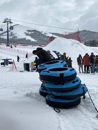 This image depicts a snow-covered ski resort environment in Yeghipatrush, Armenia, under an overcast sky. In the foreground, a stack of four blue and black inflatable snow tubing rings, each bearing the "MYLER" brand, is prominently displayed. These tubes feature black handles and tow ropes.
In the midground, a person in a dark winter jacket is bent over the uppermost snow tube. To their left, a small child in pink outerwear and a yellow hat stands near a green trash bin. Behind the stacked tubes, a group of approximately eight adults, dressed in various winter coats, stands on the snow, some appearing to observe the surroundings or interact with mobile phones. Partially visible behind this group is a promotional banner featuring a bear graphic and the "MYLER" logo. Further left in the midground, several individuals are walking on a snow-covered slope, delineated by a red safety net. A blue equipment structure is also present on the far left.
The background features snow-covered mountains and hills. A ski lift system is operational, with a tall blue pylon supporting cables from which multiple gondola cabins, some red, are suspended. Additional red safety netting is visible on the snow-covered slopes, along with a large mound of snow and a multi-story building structure on the right side of the mid-background. The sky is uniformly gray with extensive cloud cover.