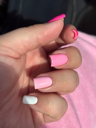 A close-up shot captures a hand adorned with a variety of colorful nail polishes. The fingertips, decorated with bright pink, soft pink, and white polishes, are arranged in a way that showcases their diverse shades and finishes. The hand is placed against a pink textured surface, possibly clothing, that complements the nail colors. The lighting suggests the image was taken outdoors, as indicated by the shadows and the bright sunlight illuminating the scene. The background is dark and blurred, focusing attention on the hand and its polished nails. The location of this scene is the Палыкавіцкі сельскі Савет, Belarus.
