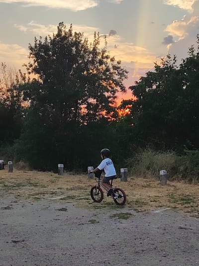 The image shows a young child riding a bicycle on a gravel path. The child is wearing a helmet and a white t-shirt. The background consists of trees and a sky with clouds. The sun is setting, casting a warm glow through the trees. The scene appears to be in an outdoor, possibly rural, setting.