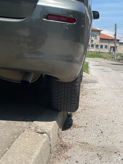 The image depicts the rear left quarter of a dark gray or greenish-gray passenger vehicle, likely a sedan or hatchback, parked partially on a concrete curb on a sunny day. The vehicle's left rear tire, displaying a winter tread pattern, is pressed against the vertical face of the curb.
The vehicle's rear bumper shows minor scratches and reflections of the surrounding environment. A red rectangular reflector or taillight element is visible on the rear left corner of the bumper. Beneath the bumper, a dark, circular exhaust pipe extends from the undercarriage. The undercarriage and inner fender well are visible and appear dirty. A side mirror, reflecting the blue sky, is partially visible on the upper right side of the vehicle.
The immediate foreground features a worn concrete curb, which is cracked and uneven in several places. Small patches of green weeds and dry vegetation grow at the base of the curb and within its cracks. The road surface adjacent to the curb is light gray, consistent with asphalt or concrete, and scattered with small pebbles and debris.
In the mid-ground, a light-colored road or path recedes into the distance. Two buildings with reddish-brown tiled roofs are prominent in the background under a clear, bright blue sky with minimal white clouds. The building on the left is a two-story structure with multiple windows, including a larger upper-floor window with white frames and an external air conditioning unit. The building to its right is a single-story structure with a visible door on its right side and fewer windows. A wooden utility pole with cross-arms and a street light fixture stands to the right of the buildings. The scene is brightly illuminated by direct sunlight, casting distinct shadows. The described location is the city of Zetiovo, Bulgaria.