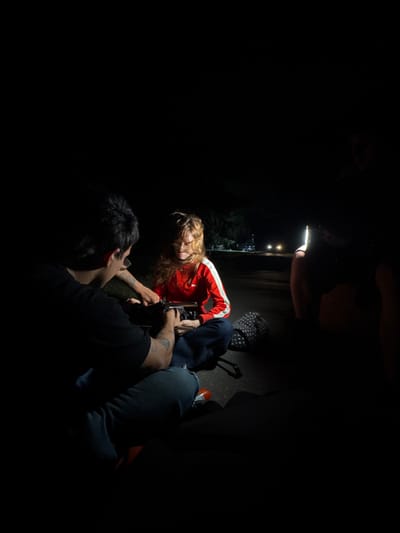 In a dimly lit, tight space, five individuals are huddled together. One shines a cell phone light on a blonde woman lying on her side in a red jacket. Two people appear to be filming her. There's a backpack in the background and the leg of another person to the right. The lighting is minimal, casting the scene in shadows and obscuring details.