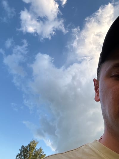 The media file is a close-up, low-angle shot of a person's head and shoulder against a bright blue sky with scattered clouds. The person appears to be wearing a dark hat and a light-colored t-shirt. Part of the person's face, including an ear and the side of their head, is visible, with some sweat glistening on the skin, suggesting a warm day. A faint glimpse of a chain necklace is visible around the neck. The background is dominated by the sky, with fluffy white clouds and some darker, more ominous clouds towards the top of the frame. A portion of a green tree with dense foliage is visible in the lower right corner. The composition is unusual, as it is taken from a very low perspective, looking upwards. The image appears to be taken outdoors, possibly in a park or open area, under daylight conditions. The time of day is likely daytime, given the brightness of the sky. The weather appears to be partly cloudy, with a mix of sun and shade. There is no visible text in the image. The orientation of the image is rotated 90 degrees counter-clockwise.