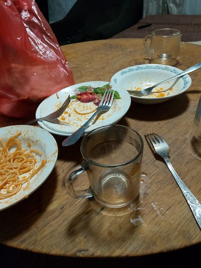 The image shows a rustic wooden table with remnants of a meal. In the foreground, a glass mug with a decorative pattern is filled with a light-colored liquid, possibly tea or coffee, and has some condensation or spills on the table around it. A silver fork with an ornate handle lies to the right of the mug. To the left, a white plate with a floral rim contains a portion of spaghetti with tomato sauce. Another white plate, further back and slightly to the right, also holds remnants of spaghetti and sauce, with a fork resting on it. A larger white bowl with a decorative blue rim is in the background, also showing signs of a meal. A crumpled red plastic bag is visible in the upper left corner of the image. The scene suggests a casual meal has recently concluded. The specific location of Кугейский, Russia, cannot be determined from the visual information provided.