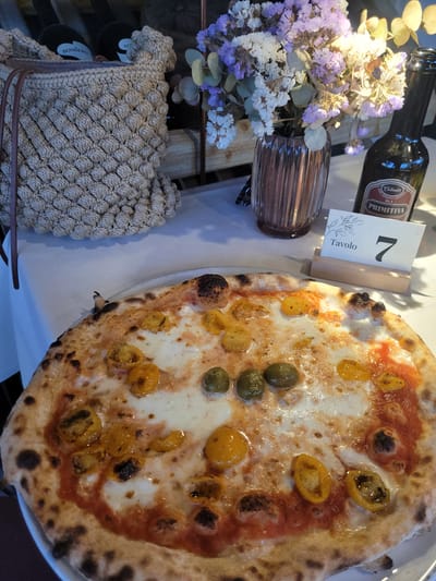 This media file captures a vibrant tabletop scene, likely in an Italian restaurant in Sogliano Cavour, Italy. The primary subject is a freshly baked Neapolitan-style pizza, prominently displayed on a white plate, occupying the right side of the frame. It features a classic puffed and slightly charred crust, melted white cheese, a rich red tomato sauce base, scattered halved yellow cherry tomatoes, and several green olives, suggesting a flavorful and authentic dish.

To the left of the pizza, a dark glass bottle is visible, its label clearly showing "Calabria" and "PRIMITIVO," indicating a bottle of Italian wine. Next to the bottle, a white table number card stands upright in a wooden holder, displaying the number "7" and the word "Tavolo" (Italian for table) written vertically, confirming a restaurant setting.

Further left, a clear, ribbed brown glass vase holds a small bouquet of dried or preserved flowers in shades of light purple and white, accompanied by green foliage. In the bottom-left corner, a textured, woven bag or basket made of natural fibers, featuring a distinctive bobble pattern, is visible. It has a label that reads "GOMBINA," and a partially visible black circular label beneath it shows "SOMIERRO."

The lighting is warm and inviting, suggesting either natural light during the day or well-executed artificial lighting, creating a pleasant dining atmosphere. The overall scene conveys the anticipation of enjoying a delicious meal in a relaxed and authentic Italian setting.