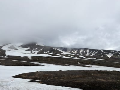 A desolate, mountainous landscape in the city of Nagornyi, Russia, is captured under an overcast sky. The terrain is a mix of dark, rocky soil and patches of snow, with larger snowfields covering the slopes of the mountains in the background. The mountains themselves are shrouded in low-lying clouds, obscuring their peaks. The scene appears barren and uninhabited, with no visible signs of human activity except for a small, red and white striped marker sticking out of the snow in the lower left quadrant of the image. The overall impression is one of a cold, remote, and rugged environment.