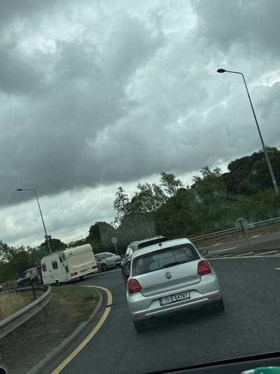 The image captures a cloudy day on a road, likely a highway, with several vehicles, including a silver Volkswagen Polo with license plate 171-D-64787, and a white caravan. The scene is set in or near Slieverue, Ireland, as indicated by the license plate format and context. The road appears to be curving, with a guardrail and lane markings visible. Trees and foliage line the sides of the road, suggesting a semi-rural or suburban environment. A street lamp is present, and a speed limit sign displaying "80" is visible in the distance. The sky is overcast with heavy, dark clouds, hinting at potential rain. The vehicles are stopped or moving slowly, possibly indicating traffic congestion. No individuals are clearly visible within the vehicles.