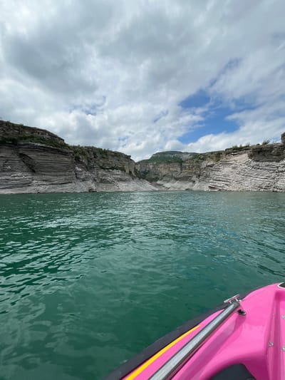 The image depicts a scene in Chirkey, Russia, showing a body of water with rocky cliffs on one side and a pink boat on the other. The water appears calm, with visible ripples and a green hue. The cliffs are gray and layered, with some vegetation growing on them. The sky is cloudy with patches of blue. A section of a pink boat is visible in the upper right corner of the frame.