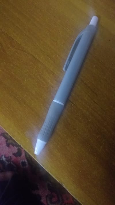 The image shows a close-up, slightly blurry view of a gray pen lying on a wooden surface. The pen has a white tip and a clip on the side. The wooden surface has a visible grain and appears to be a table or desk. In the lower right corner, a portion of a patterned rug with red and dark colors is visible, suggesting the setting is indoors. The lighting appears to be artificial, casting a slight glare on the pen's tip. There is no activity or event depicted, only the still object. No people are visible in the frame. There is no visible text in the image. The location context provided is Ghafurov, Tajikistan.