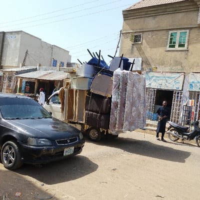 This image depicts a street scene in Dan Maliki, Nigeria. A white, light-duty truck is centrally positioned, heavily loaded with an assortment of household furniture, including multiple wrapped mattresses, stackable sofa cushions, chairs, and a wooden cabinet. The cargo is visibly secured to the truck bed, extending beyond its physical dimensions.
In the foreground, a black Honda sedan with a Nigerian license plate "SW-520HA" is parked on the left side of the unpaved road. To the left of the loaded truck, a person wearing a white garment and a red cap is partially visible near the vehicle's front.
On the right side of the frame, a man dressed in a dark blue or black traditional garment stands beside a parked black motorcycle. He is positioned in front of a multi-story building constructed from beige masonry, featuring green-framed windows on its upper floor. The ground level of this building appears to house commercial establishments with metal grates covering their entrances.
In the background, on the left, a larger, light-colored building with "PLAZA" prominently displayed is visible, alongside smaller structures with corrugated metal roofs that resemble market stalls or shops. Overhead utility lines span across the scene. The road surface is predominantly unpaved dirt, and the environment is illuminated by bright daylight, casting distinct shadows.