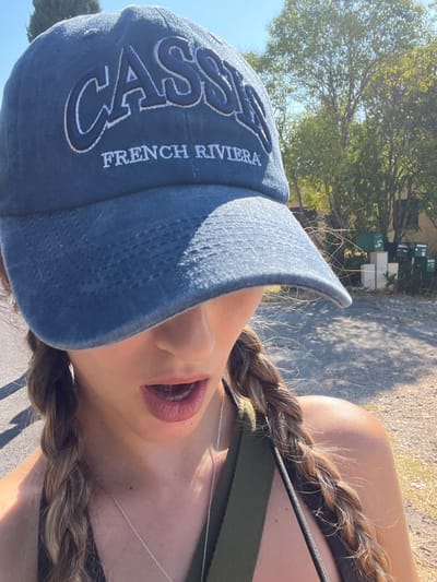 This close-up outdoor photograph depicts an individual in a bright, sunny environment in Puyloubier, France. The subject, a female, is partially visible, with the view extending from just above the nose to the upper torso. She is wearing a faded blue baseball cap with "CASSIS" embroidered in large white letters and "FRENCH RIVIERA" in smaller white letters on the front; the cap's brim shades her upper face. Her mouth is slightly open, revealing pink lips. Her light brown hair is styled in two visible braids, resting over her shoulders. She wears a dark top, possibly a tank top, with a prominent olive green strap visible on her right shoulder. A thin silver chain necklace is also visible. The surrounding environment consists of a light-colored gravel or dirt ground in the foreground. In the background, under a clear blue sky, are green trees with dense foliage. To the right, a cluster of dark green mailboxes or utility boxes are mounted on lighter, cubic pedestals, indicative of a roadside or semi-rural setting. No specific actions or interactions beyond posing for the photograph are discernible.