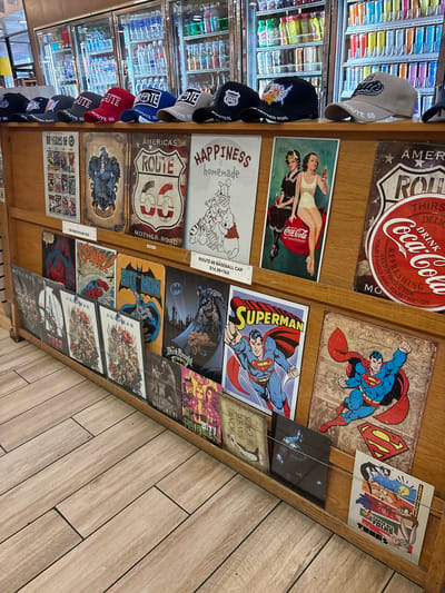 This media file is a close-up, eye-level shot of a display shelf filled with merchandise. The items on display include several baseball caps with "Route 66" branding and various graphic designs, as well as several tin signs featuring iconic pop culture imagery. These include signs for Coca-Cola, Superman, Batman, and Looney Tunes characters. Some signs also reference "Route 66" and "Mother Road." There are also comic book style panels displayed. The background shows shelves stocked with bottled drinks in a cooler. The overall setting appears to be a retail environment, possibly a gas station or convenience store, given the presence of the drink cooler and the Route 66 memorabilia, which evokes a classic American roadside theme. The lighting suggests it's daytime.