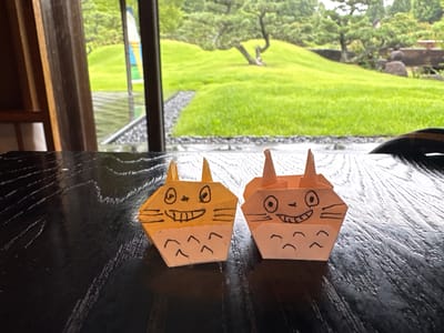 Two origami figures, resembling characters from the animated film My Neighbor Totoro, sit on a dark, glossy wooden table. The figure on the left is predominantly yellow with black drawn features of eyes, a wide smile, whiskers, and a belly pattern. The figure on the right is orange with similar black drawn features. In the background, a lush green lawn slopes upwards, with a gnarled tree visible on the hill. Beyond the lawn, a glass window or door reveals a rainy, outdoor environment with reflections on the wet ground. The scene appears to be set in a garden setting, possibly at the Ghibli Park in Nagakute, Japan, given the Totoro-themed origami.