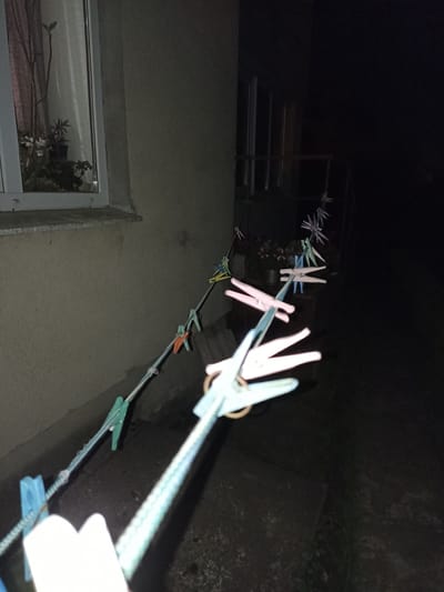 In Pochaiv, Ukraine, a clothesline is strung across a dark outdoor area, likely a courtyard or alleyway, adjacent to a textured, beige wall. The clothesline is adorned with numerous colorful clothespins, including pastel pink, blue, and green, some of which appear to be plastic and others possibly wooden. In the background, a window with a plant is visible, and beyond that, the scene transitions to darkness. The image is taken at night, as indicated by the lack of ambient light and the artificial illumination on the clothespins in the foreground, suggesting a flashlight or camera flash. The perspective is low, looking up towards the clothesline, with the foreground dominated by the prominent clothespins and the textured rope of the clothesline.