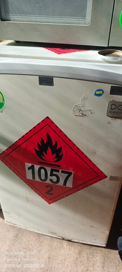 This image shows a close-up of a white appliance, possibly a refrigerator or washing machine, with several stickers and labels. The main focus is a large, diamond-shaped hazard symbol with a red border and a black flame icon, indicating flammability. Inside the diamond, the numbers "1057" and "2" are clearly visible. There are also smaller rectangular labels with text, some of which appear to be expiration dates, with dates like "18.02.2023" and "16.02.2024" visible. A sticker with the "DAEWOO ELECTRONICS" logo is present on the right side. A whimsical sticker of a person in a contorted pose is on the upper right. Another green sticker with the word "th" is visible on the left. The appliance itself appears somewhat aged and dusty, suggesting it is in use or has been stored for some time. The bottom of the image includes a timestamp and location information: "2026/06/30 17:32", "Гурьевск, Россия" (Guryevsk, Russia), and "Infinix ZERO 30". The lighting suggests it is daytime. The overall scene is a still life of an appliance with various markings.