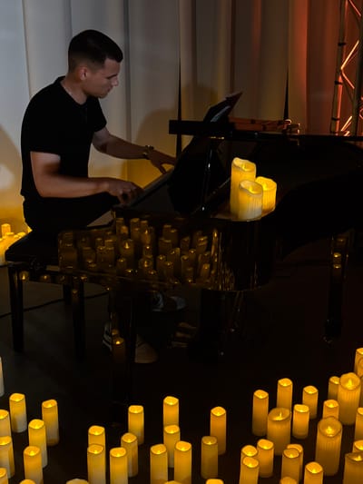 A man wearing a black t-shirt and black pants plays a black grand piano, surrounded by numerous flickering LED candles. The candles are placed on the floor around the piano, on the piano itself, and on the piano bench. The room appears to be dimly lit, with the warm glow of the candles illuminating the scene. In the background, a beige curtain and part of a stage lighting rig are visible. The man is focused on the piano keys, with his hands positioned to play. There is no visible indication of the location being in Ilyanskiy selskiy Sovet, Belarus, nor is there any evidence to suggest a forensic context.