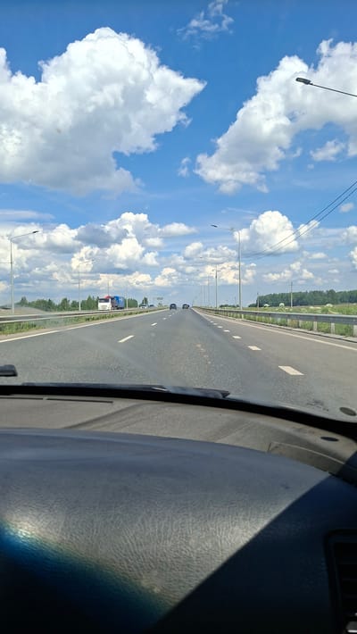 The image depicts a view from inside a vehicle traveling on a highway. The perspective is from the passenger seat, looking out the window towards the road and the sky. The road is asphalt and appears to be a multi-lane highway, with white lane markings visible. Several vehicles, including cars and trucks, are seen driving ahead on the highway.
The sky is a vibrant blue with numerous fluffy white cumulus clouds scattered across it. The sun is shining, indicating a clear and bright day, likely daytime. The presence of streetlights lining the highway suggests it's a well-maintained road, possibly in a developed area or a highway outside a city. Green trees line the side of the road in the distance, and a metal guardrail separates the highway from the embankment.
The interior of the vehicle is partially visible on the right side of the frame, showing a textured dashboard with a shadow cast across it, likely from the sun. The orientation of the image is rotated 90 degrees counter-clockwise, making the sky appear on the left and the road stretching into the distance on the right. There are no visible people in the frame, and no text is discernible. The overall scene conveys a sense of travel and movement on a sunny day.