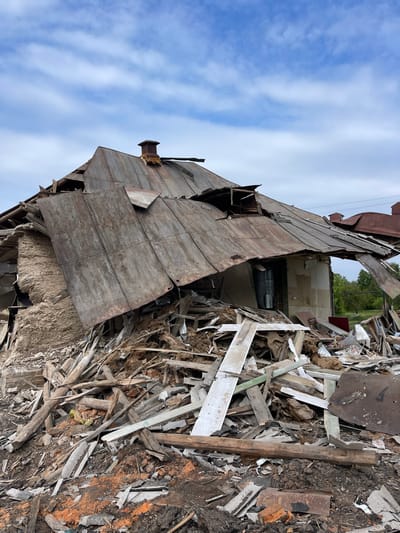 The image shows a destroyed building in Bilopillia, Ukraine (latitude: 51.139, longitude: 34.285). The building is a single-story structure with a wooden roof that has collapsed. The roof is lying on the ground, partially covering a pile of debris. The debris consists of wood, metal, and other building materials. There are no people in the image. The sky is blue with some white clouds. The scene is located in a residential area, as evidenced by the presence of other buildings in the background. The scene suggests that the building was destroyed by an explosion or other violent event.
