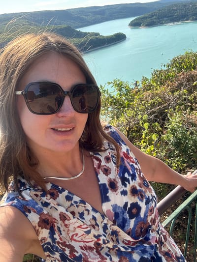 A woman with brown hair, wearing sunglasses, a silver necklace, and a floral print dress, poses for a selfie outdoors. She is looking towards the camera and smiling. The background shows a body of water, possibly a lake, surrounded by green hills. The scene is likely located at the Lac de Vouglans, a reservoir in the Jura department of France, near Moirans-en-Montagne. The sky is clear and sunny.