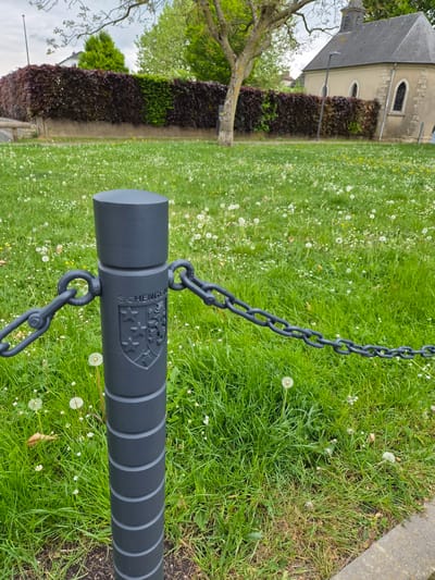 The image shows a close-up of a black metal bollard connected to a chain, standing in a grassy area. The bollard has the word "SCHENGEN" and a coat of arms embossed on its side. The background includes green grass with dandelions, a hedge of burgundy foliage, and a building with a gray roof. The scene is likely located in the city of Schengen, Luxembourg, based on the inscription on the bollard.